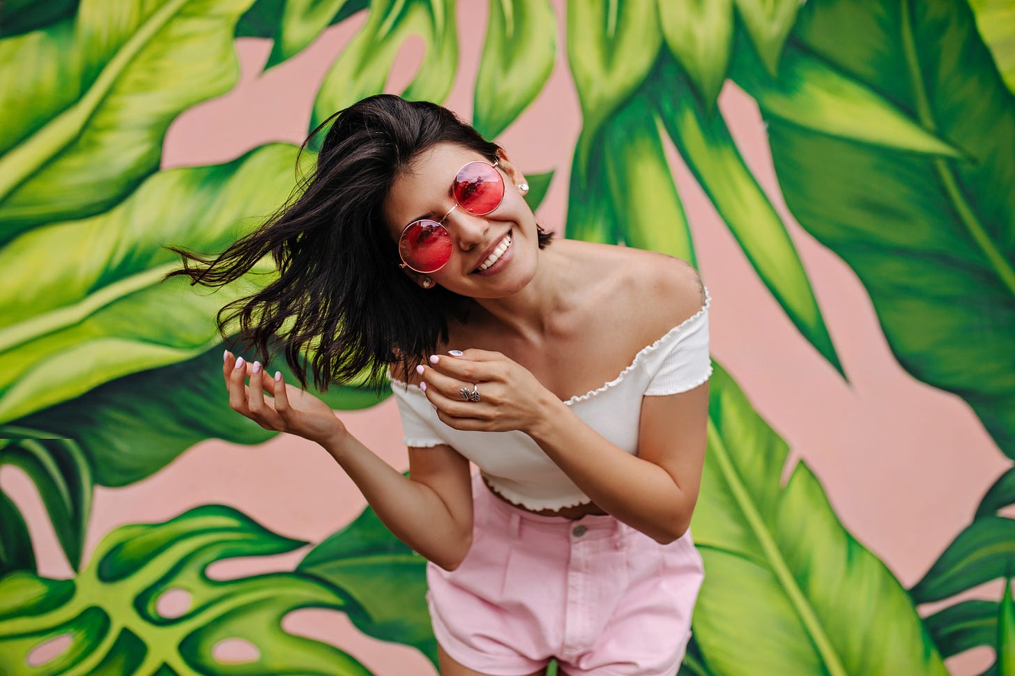 A woman in bright colors (and dark hair) smiles and poses against a painted pink and green background.
