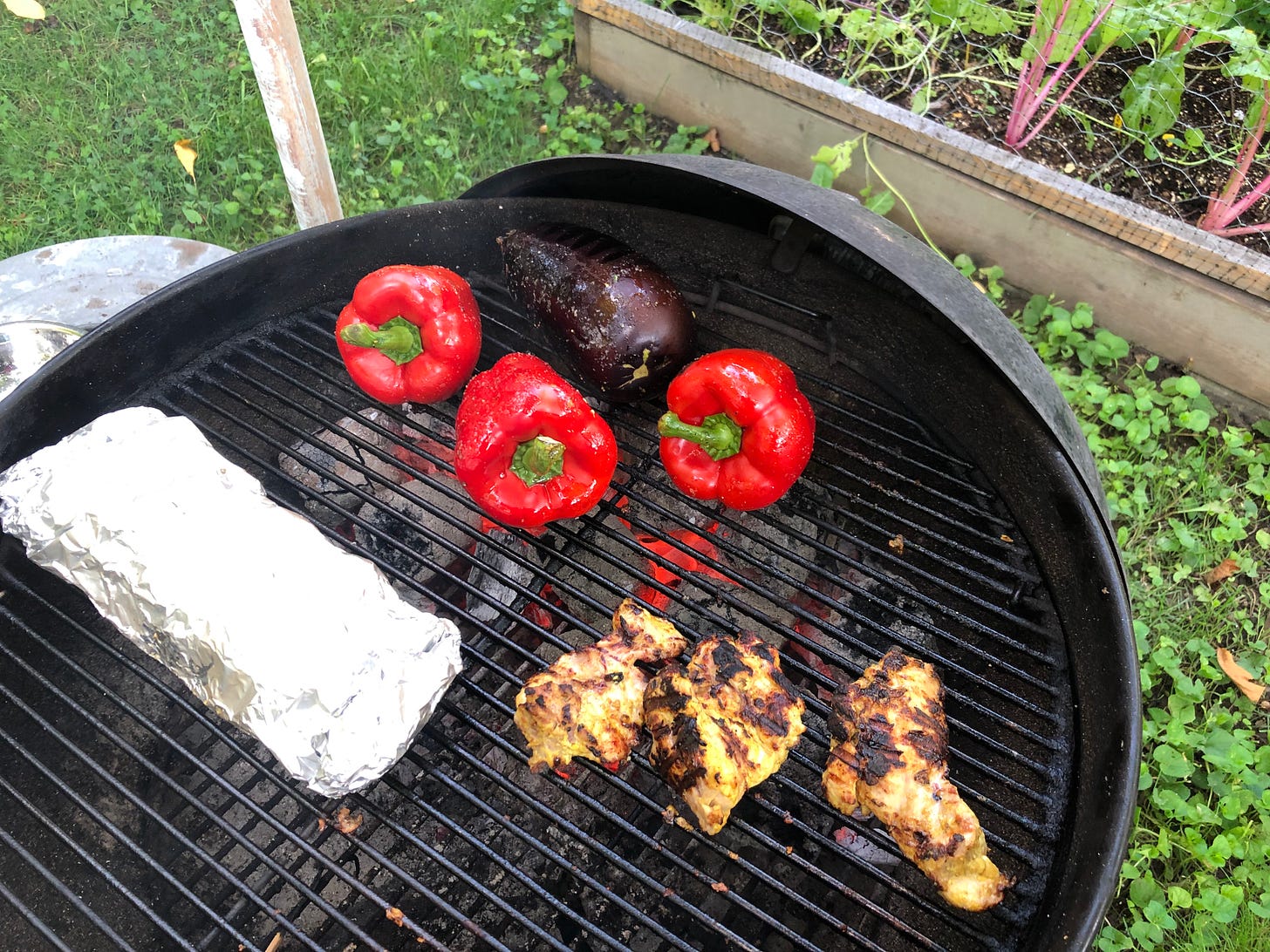 A charcoal grill with XYLO charcoal blocks burning. Three red peppers and an eggplant are directly over the coals. A foil packet of potatoes and some marinated chicken thighs are offset slightly and not directly over the coals.