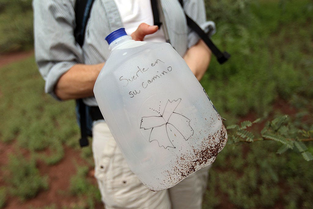 A person wearing a striped button down and khakis holds up a gallon water jug that has a marker drawing of a butterfly on it with the words "Suerte en su camino," or "Good luck on your journey"