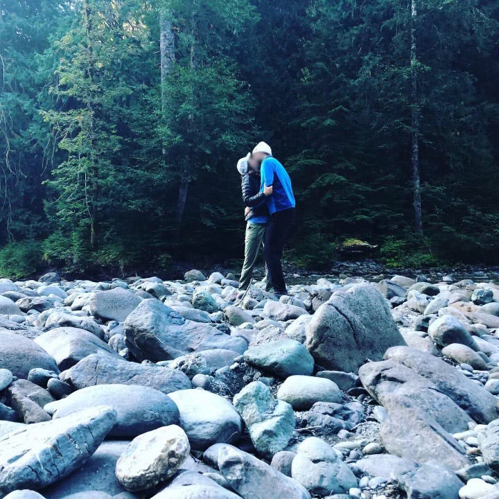 A white woman and man embrace and kiss on a bare rocky creekbed, the forest looming behind them