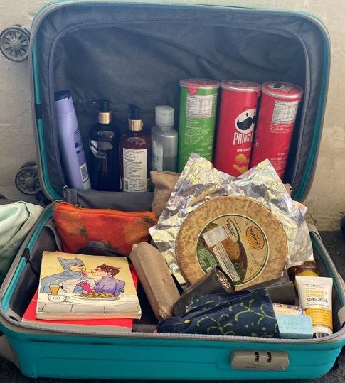 image of a hard cabbin luggage size suitcase opened up to be used as a shelf. The top has vertically standing toiletories and pringles boxes and the bottom has khakhra, pouches and books