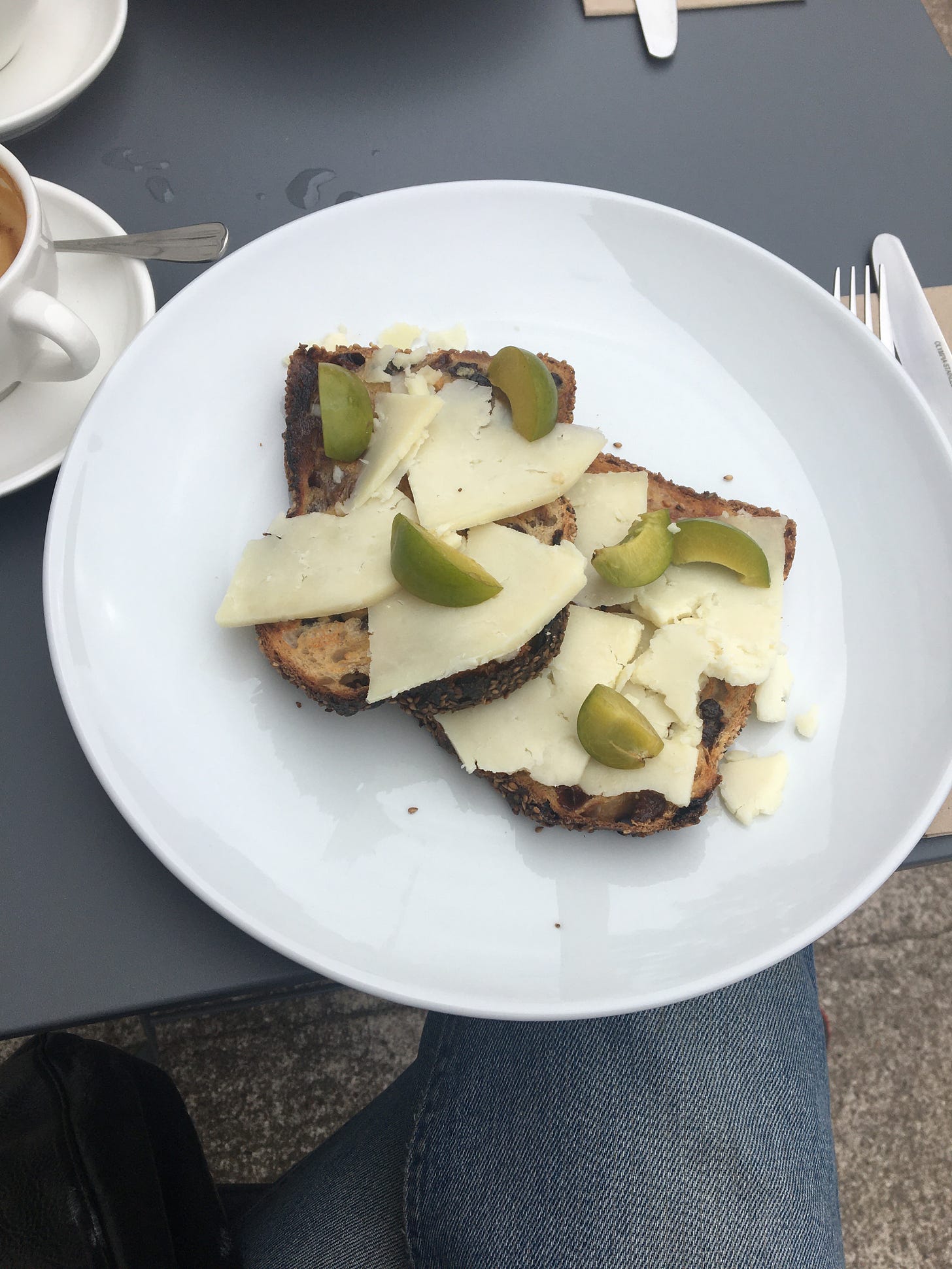 An outdoor photo of white modern crockery against a grey metal table, Kate's jeans underneath. There is some cutlery to the right, and a cup of drunk coffee cut off by the frame. On the central plate are two slices of toast, with a pale hard cheese with green plum-like segments dotted over. 