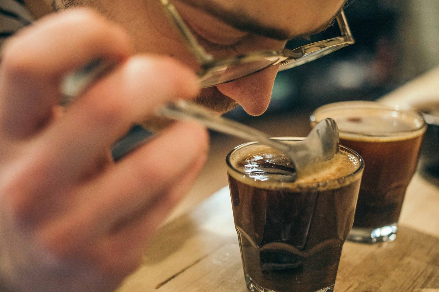 A close up image of a white man with glasses using a spoon dipped into a small bowl of coffee close to his nose while participating in a traditional coffee cupping tasting exercise.