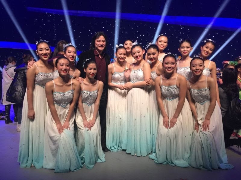 thirteen asian girls in long blue and white dresses smile at the camera onstage; their dance teacher stands with them.