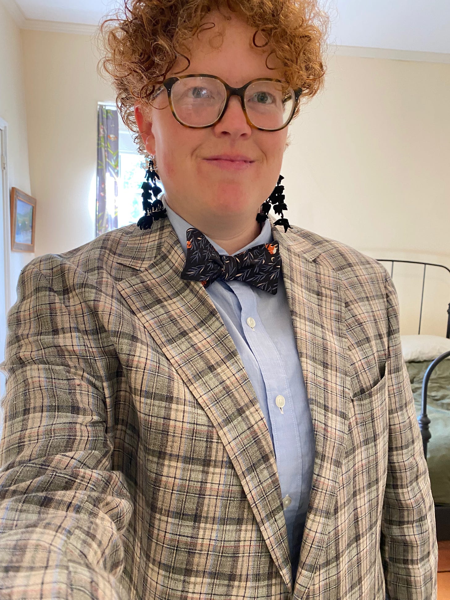 I am standing in a bedroom, wearing a blue button-down shirt and a green, brown, and pink patterned jacked. I have on long dangly black earrings made of folded pieces of fabric, and a bowtie patterned with foxes and small grasses. I have short red curly hair and big glasses, and I am smiling slightly.