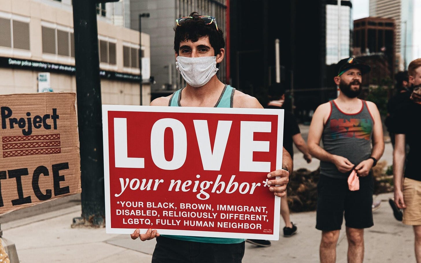 A person is holding a sign that says: LOVE your neighbour* *your black, brown, immigrant, disabled, religiously different, lgbtq, fully human neighbour