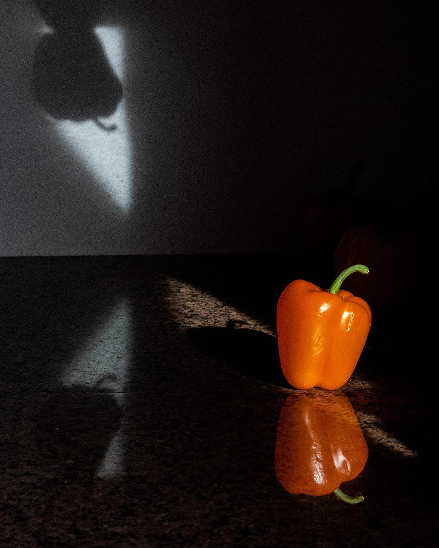 An orange bell pepper is photographed on a kitchen countertop. Sunlight causes the pepper to be reflected in the countertop and multiple shadows of the pepper to be created in the scene. 