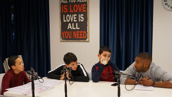 foto van drie jongens en een man die aan een tafel zitten met daarop statieven met microfoons. Op tafel liggen papieren en er hangt een poster met daarop de tekst love is all you need