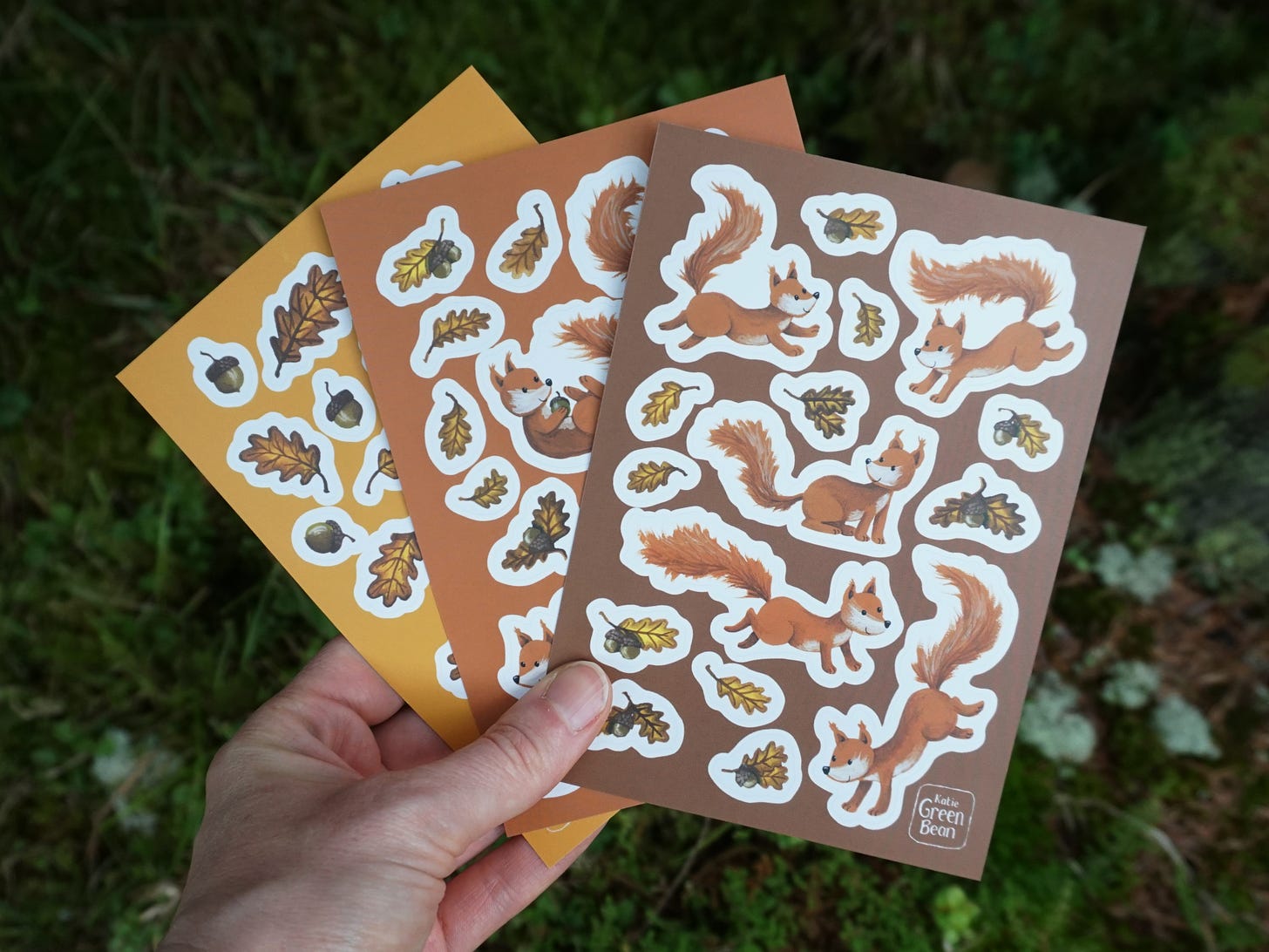 The same hand holds a fan of three more sticker sheets, this time with playful red squirrels, acorns and oak leaves. 