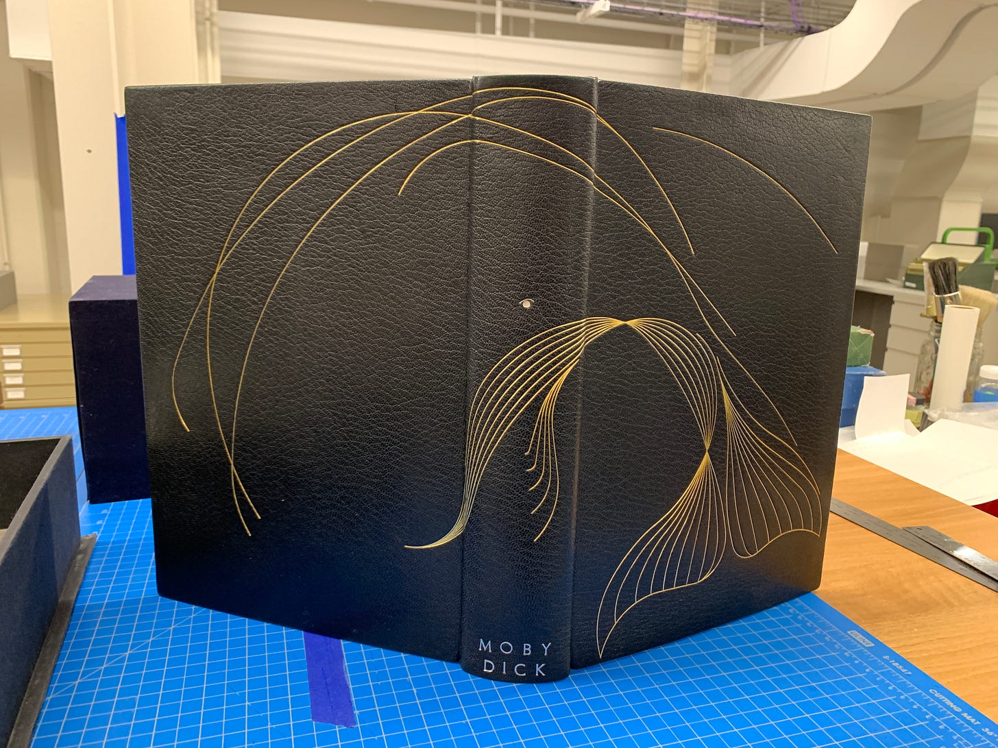 Glaister's binding of Moby Dick. The main binding is black leather, with gold tooling in the shape of a whale that crosses both back and front cover. The Whale's eye is a silver tool dot one-third of the way down the spine. 