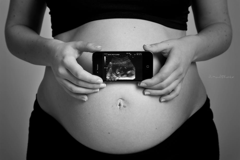 a very pregnant woman wearing a crop top and pants with her bare belly exposed holding a picture of an ultrasound of her fetus