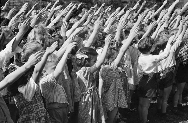 Several dozen young elementary-age children raise their arms toward a flag in an outdoor setting