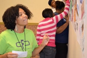 This is image is of a light Brown Black woman with thick curly hair. She is biting her bottom lip and looking at a paper on a wall and adding a post it note. She is wearing a lime green shirt with purple writing that says CFC Crunk Feminist Collective. She is the younger version of the same woman in the next image.