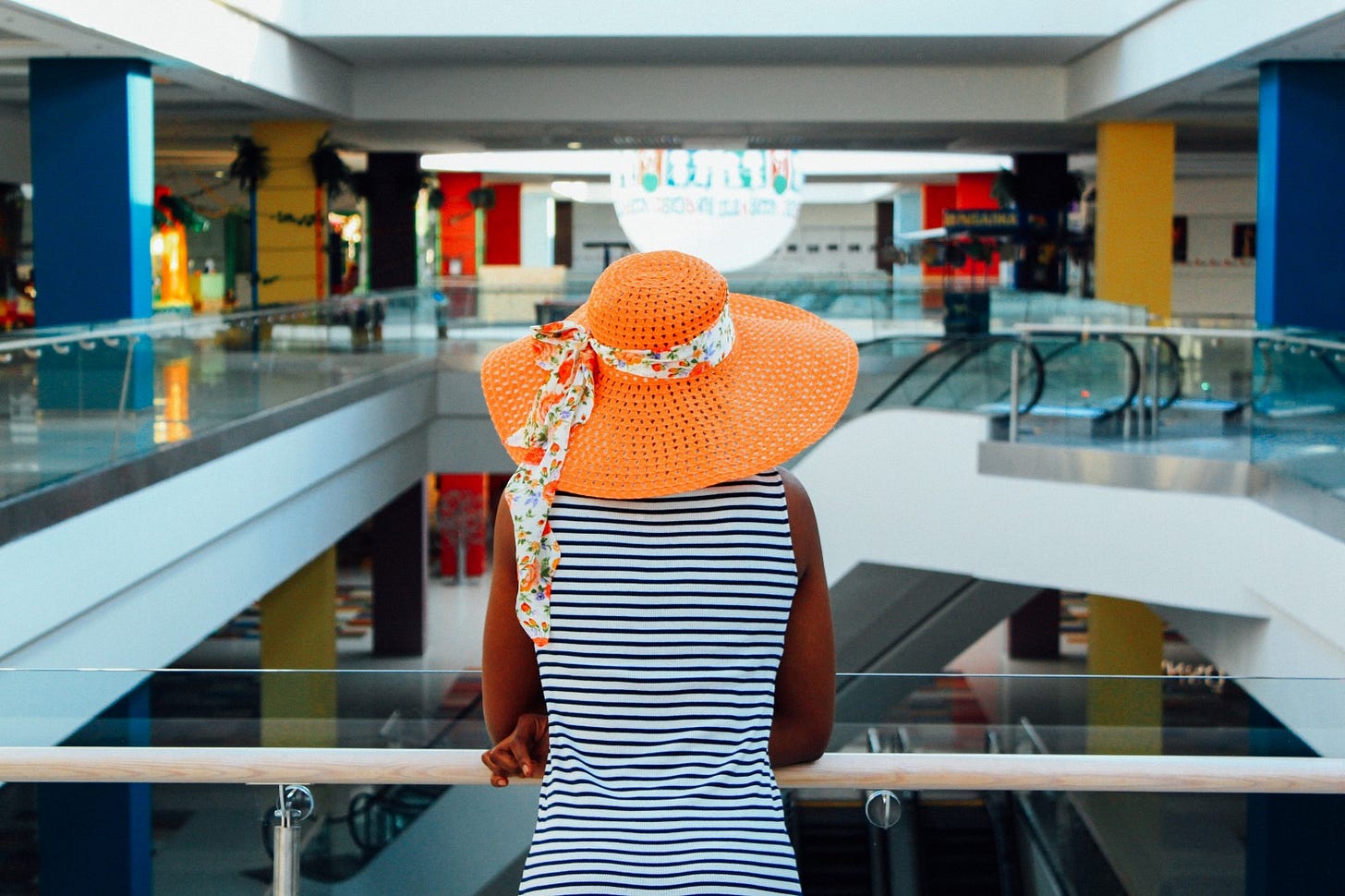 A woman has her back to the viewer. She is wearing a blue and white striped tank top and a large orange sunhat. She is a shopping mall.