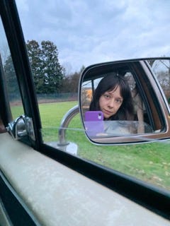 A white woman with dark brown hair is seen in a reflection of a side mirror of a truck. Green grass is behind her image.