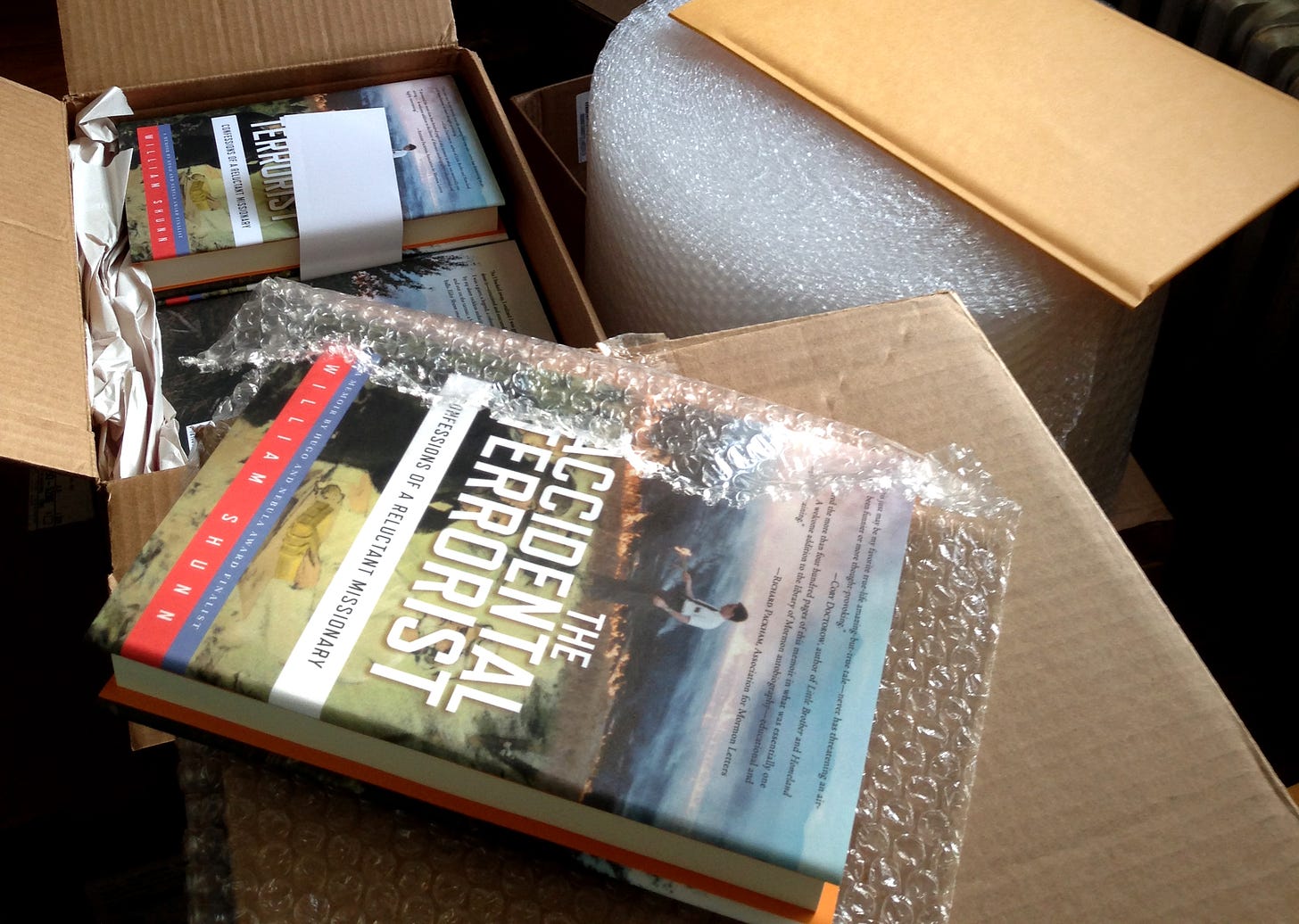 A hardcover copy of The Accidental Terrorist half-wrapped in bubble wrap sits next to an open carton of more copies of the book, with mailing supplies nearby.