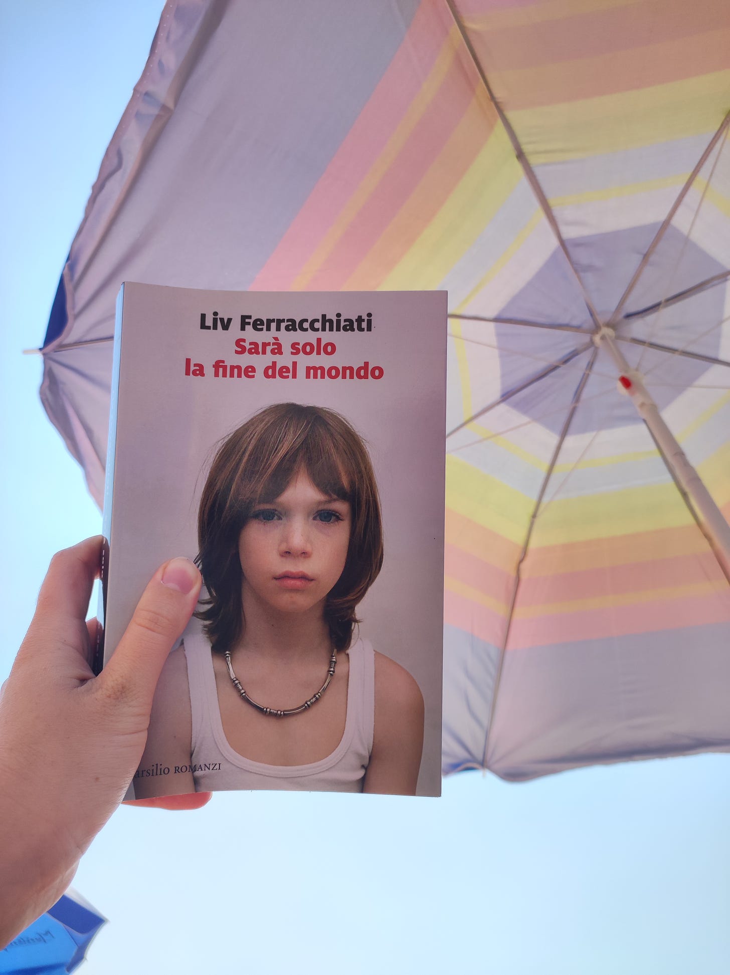 Una mano tiene un libro. Si intitola "Sarà solo la fine del mondo" ed è di Liv Ferracchiati. Sulla copertina c'è una bambina in canuttiera. Sullo sfondo della foto c'è un ombrellone a righe colorate e si intravede un cielo azzurro.