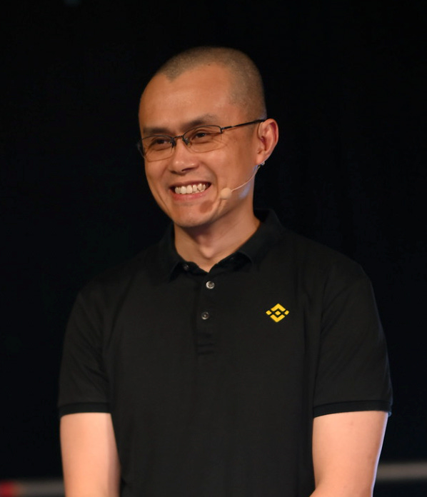 Changpeng Zhao, smiling. He's wearing a black polo shirt with a small yellow Binance logo on the chest, and a Madonna microphone.