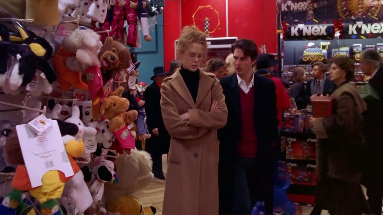 Film still from Eyes Wide Shut. Nicole Kidman and Tom Cruise stand in a toy section in a busy mall. Nicole has her arms crossed, looking at the ground.