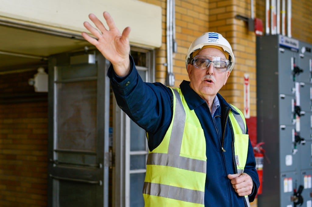 In a hard hat and bright yellow safety vest, Bob Bonnett gestures off to the distance as he looks toward and past the camera. He stands in an industrial setting, the inner workings of our Easterly wastewater treatment plant.