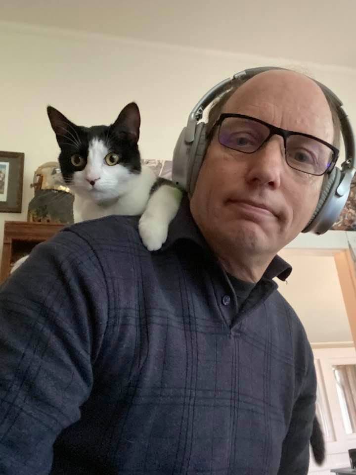 Author Brian R Price in the office wearing headphones, black and white cat perched on his shoulder