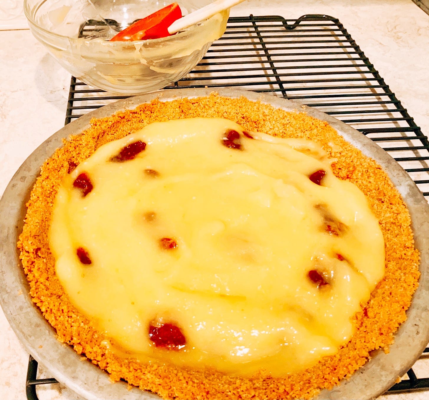 A lemon curd pie in a graham cracker crust is dotted with berry preserves. It sits on a wire rack. Behind it, a glass bowl that previously held the lemon curd now only hold a red spatula.