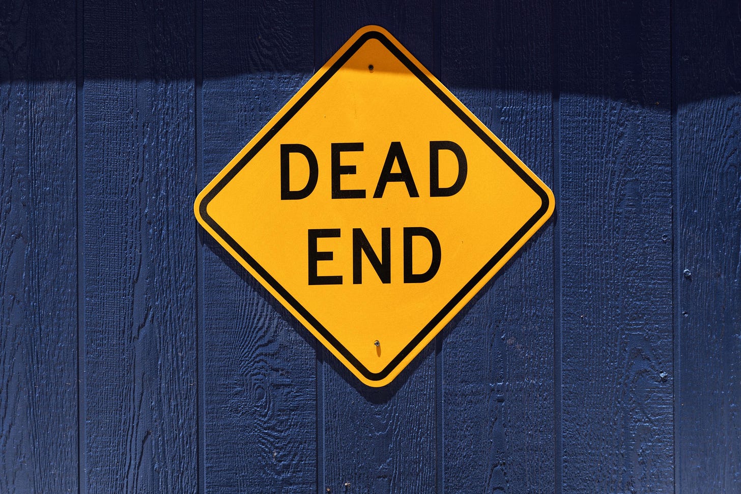 Image of a yellow sign that says “ead end” mounted on a blue wall, for article by Larry G. Maguire