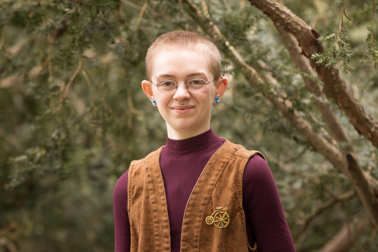 A photo of Cavar, a white, genderless person, standing among trees. Cavar has glasses and visible eyebrow, ear, septum, and philtrum piercings. They are wearing a wine-colored mock neck shirt underneath a light-brown vest with a vintage bicycle brooch.