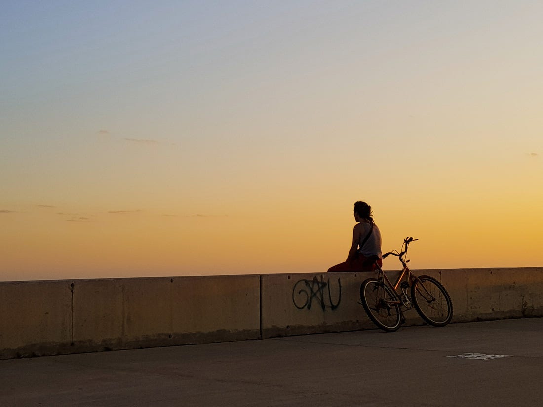 Foto colorida de um pôr do sol alaranjado. Sobre uma mureta, uma moça observa a paisagem, de costas para a câmera. ao lado dela está sua bicicleta
