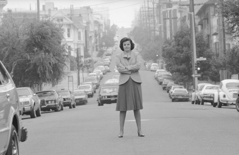 Dianne Feinstein, standing astride a San Francisco street in a skirt, jacket and bow around her neck in 1981. Parked cars line both sides of the street, and a car is approaching from behind in the distance.