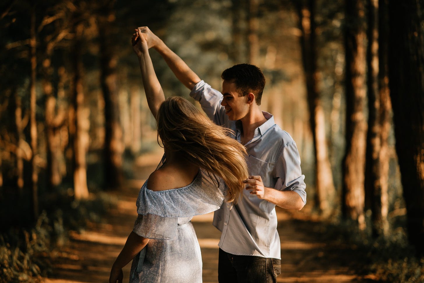 A man and a woman dance in the woods. The man is spinning the woman and smiling. The woman’s hair is drifting in the wind.