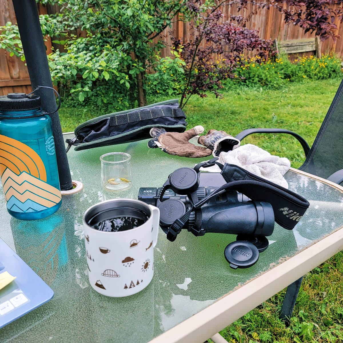 a glass, outdoor table filled with an array of items. a nalgene waterbottle, well loved gardening gloves, binoculars, seeds in a jar, a towel, a cup of coffee, and the edge of a laptop