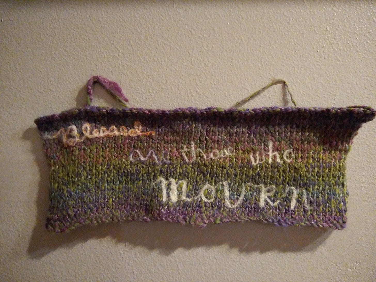 A longer piece of knitting hanging on a wall, with the words "Blessed are those who MOURN" felted onto it