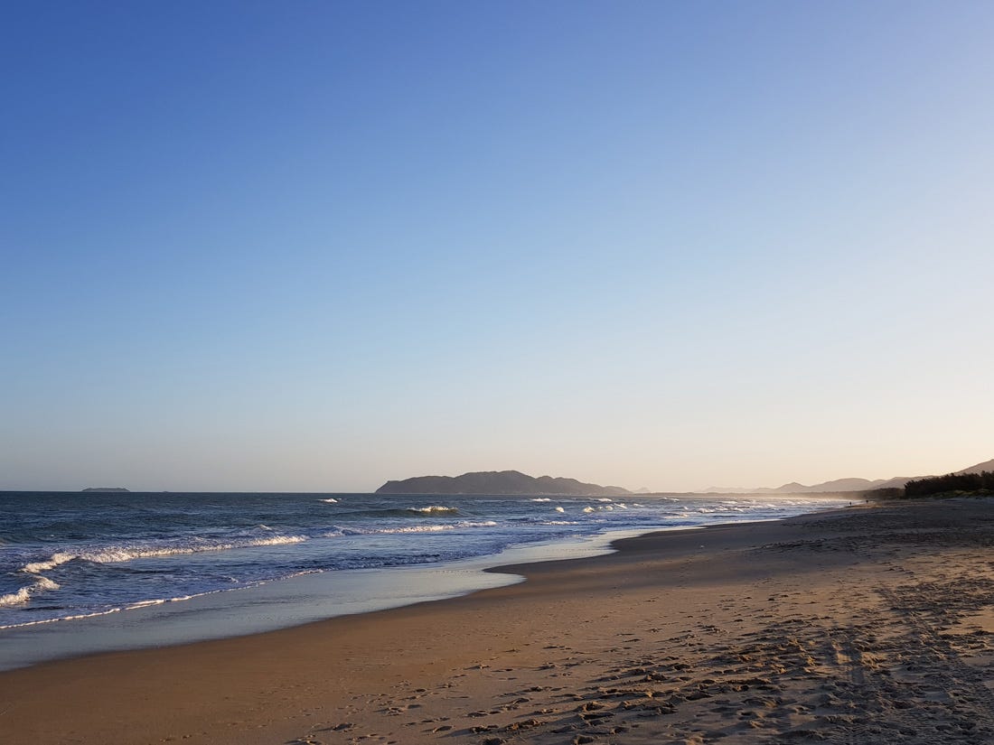 Foto colorida de um pôr do sol na praia. À esquerda da imagem está o mar, à direita está a areia alaranjada pela luz da hora dourada. O céu está azul sem qualquer nuvem.