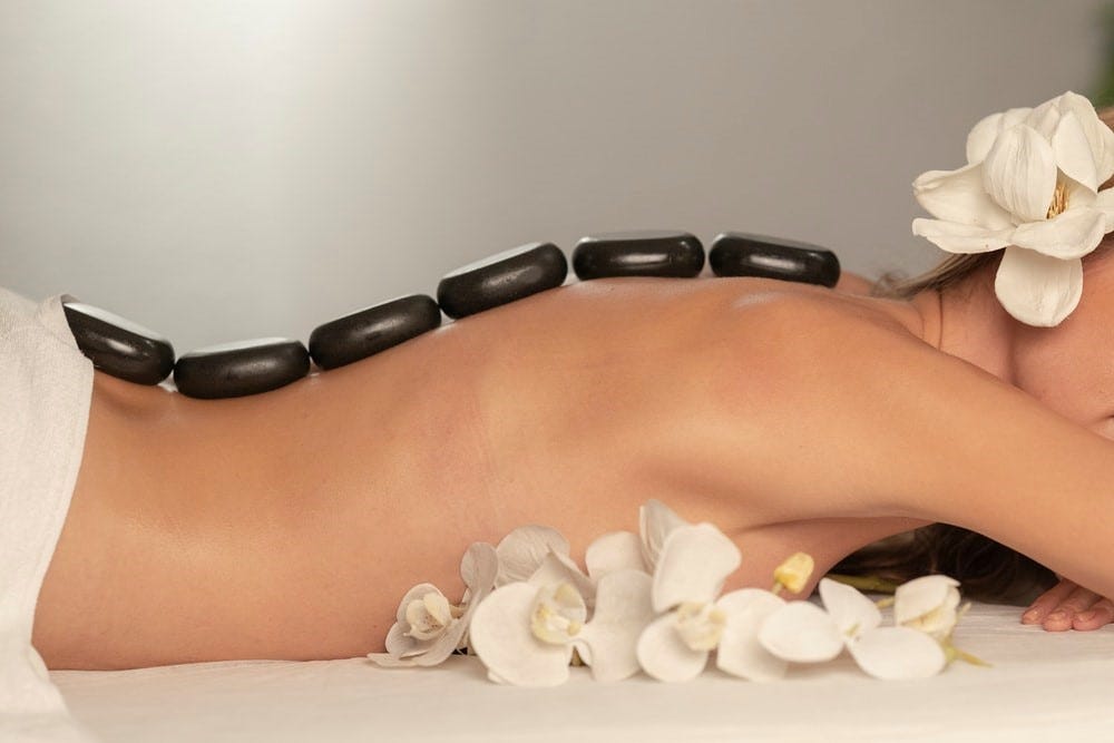 A row of stones placed along the spine of a woman lying face down wearing a towel around her waist and an exotic flower in her hair.
