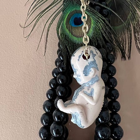 A still image of a plastic fetus with a hole cut through its head and a chain running through it, hanging against a string of black beads and a peacock feather earring