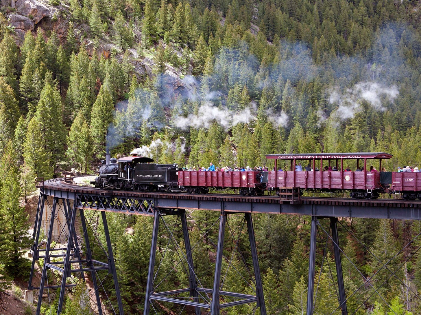 An old fashioned-looking train travels along a mountainous track surrounded by pines.