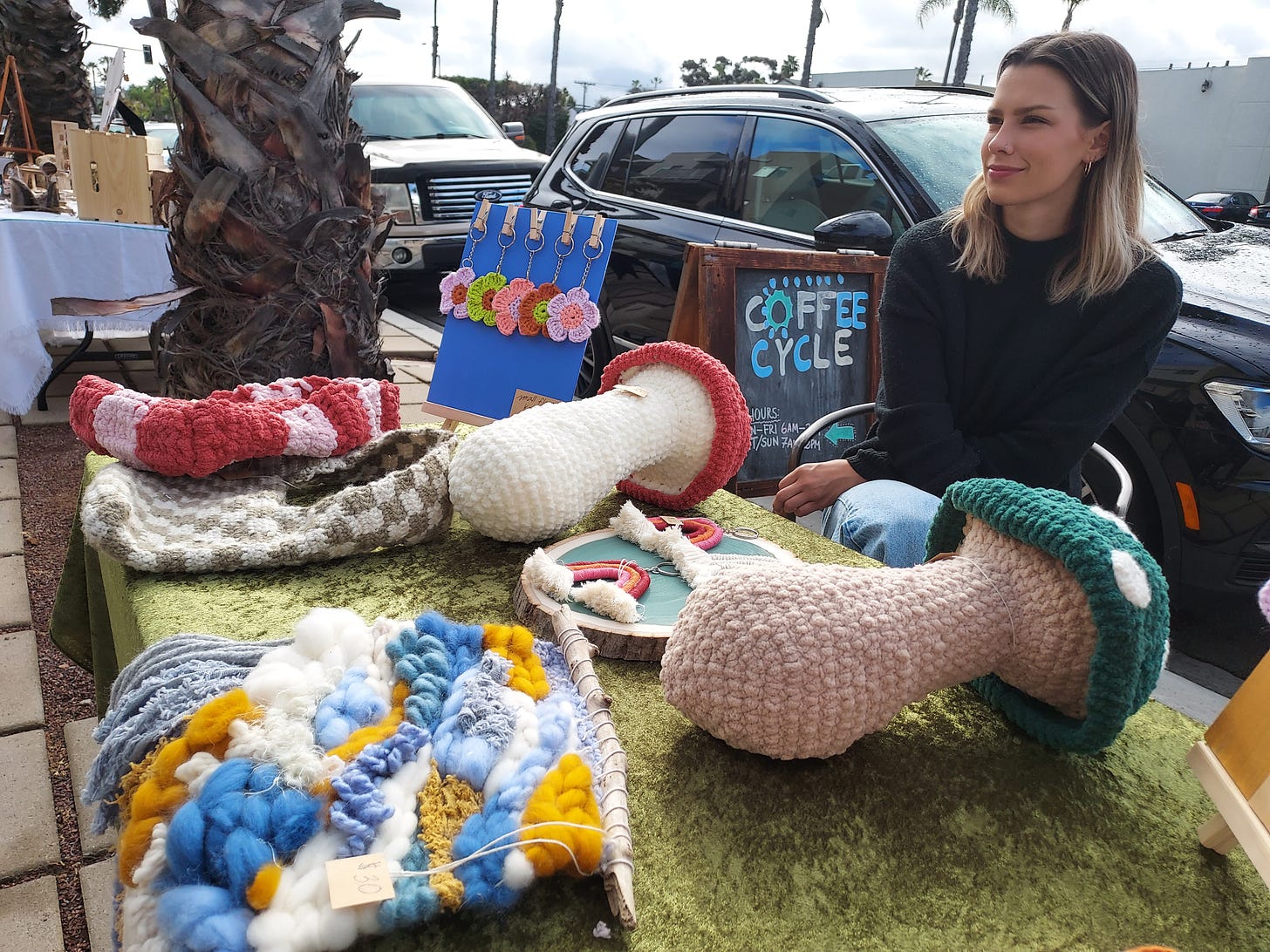 A woman looks into the distance sitting behind her table at a local makers market. The table is covered with weaved goods like oversized mushrooms, purses, and keychains.