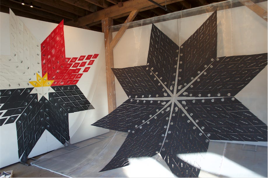 Two large quilt-like stars constructed of wired-together diamonds with words and images of animals on them are in progress of being hung in a gallary.