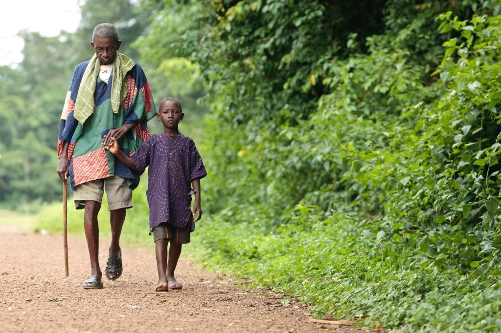 A boy helps Samuel Nicol (age unknown), who was blinded by river blindness, walk through the village of Gbonjeima, Sierra Leone, on Saturday, July 14, 2012. The Task Force for Global Health is working with partners to eliminate river blindness by 2025. Copyright: Olivier Asselin, courtesy of the Global Network for Neglected Tropical Diseases.