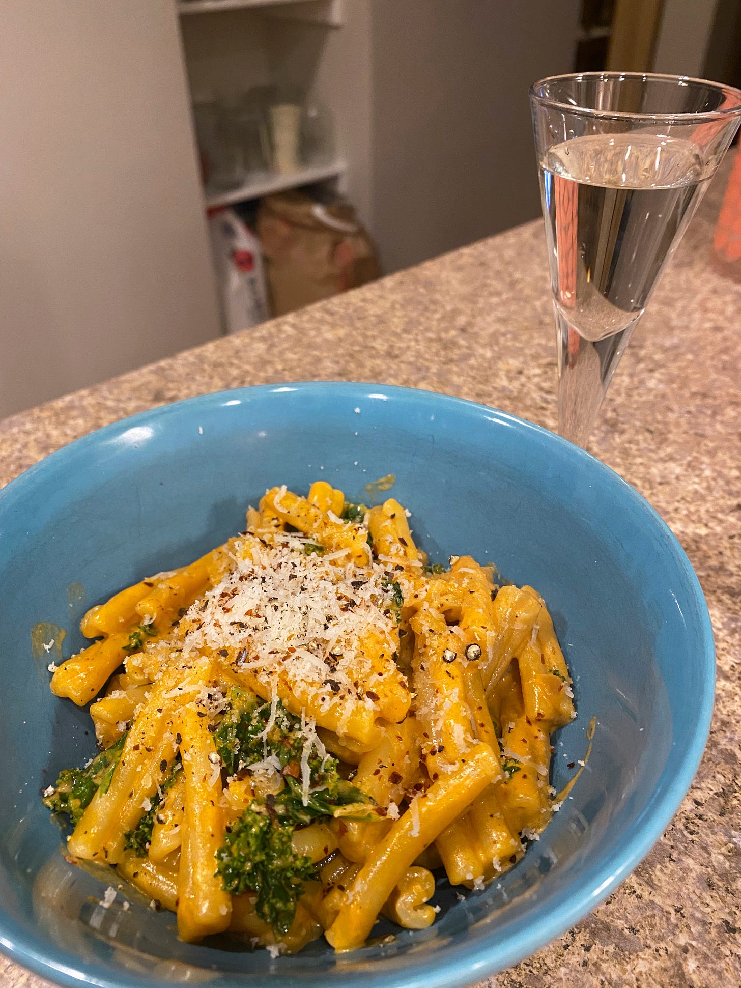 A blue bowl of casarecce in vodka sauce with pieces of green kale, and grated parmesan and chili flakes on top. Beside it is a small glass of white amaro.