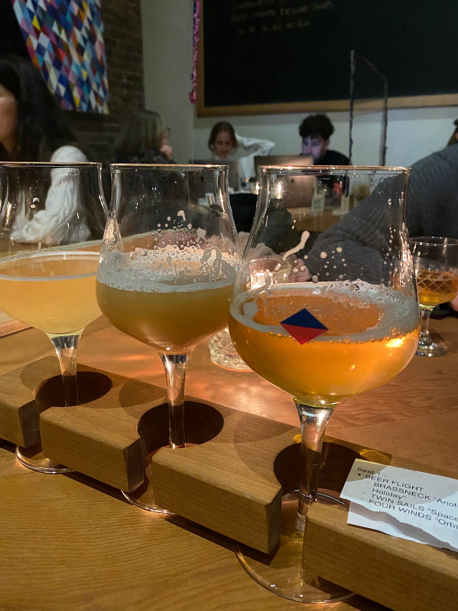 A flight of three beers in tulip glasses. A red and black diamond logo is visible on the front of the nearest glass.