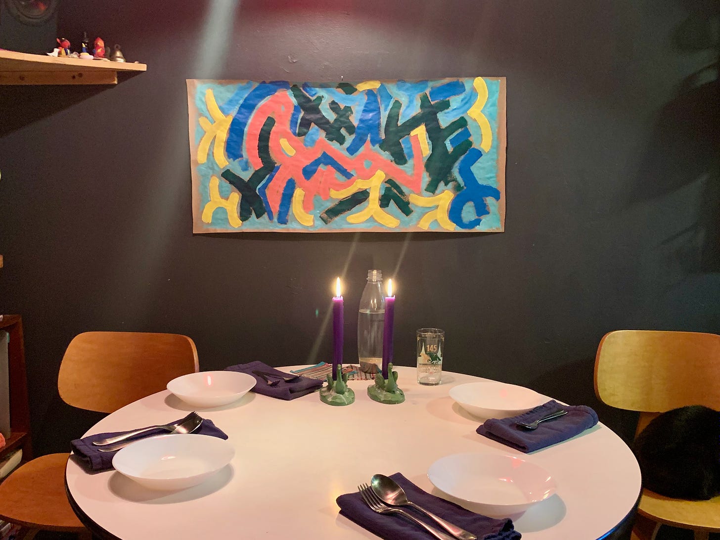 A round kitchen table, set for dinner, two blue candles are burning, above the table a painting with colors like, peach, royal blue, light blue, yellow - dance in abstraction.