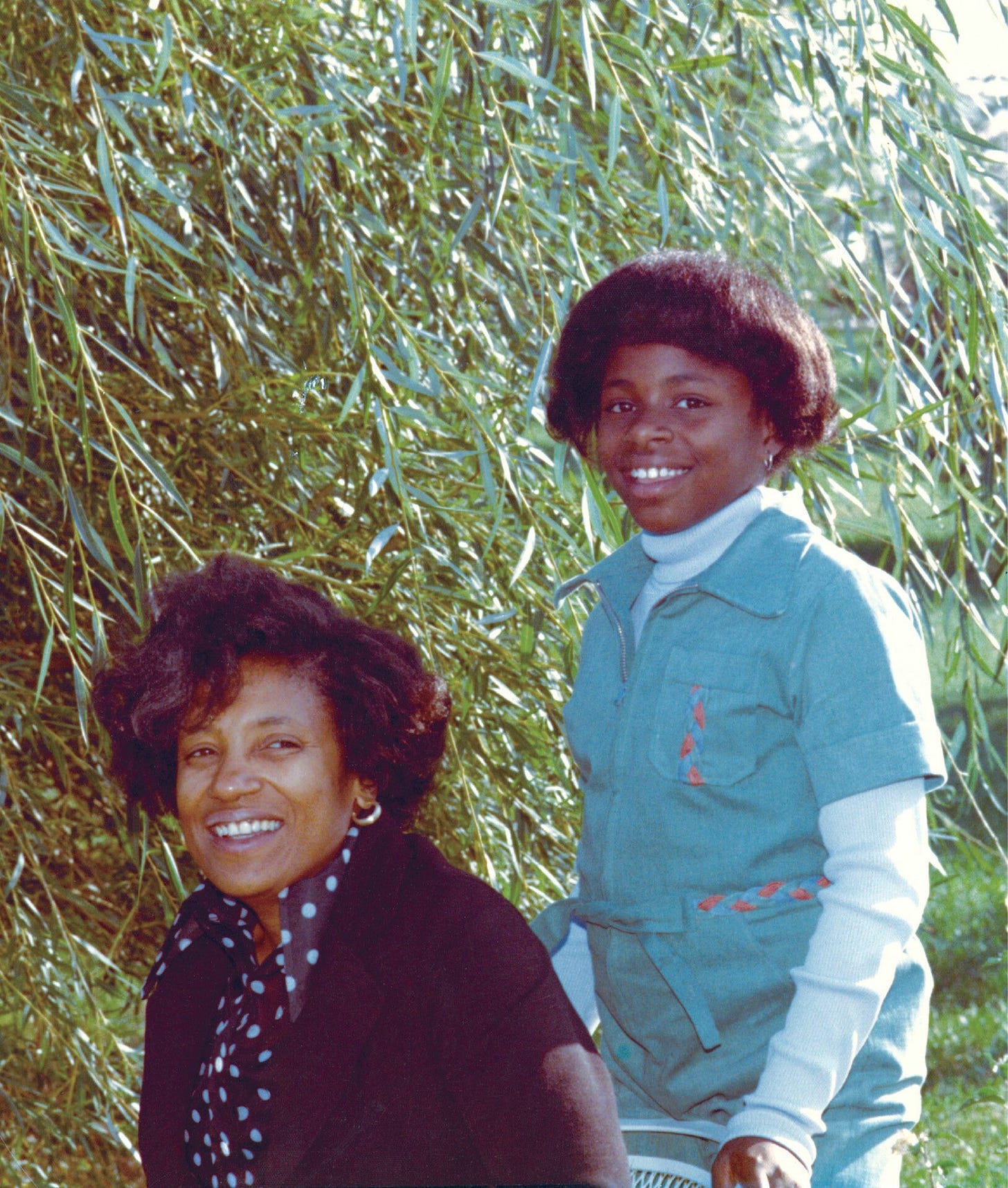 a woman kneeling with a girl standing tall next to her. a tree is in the background. both are afro-descended.