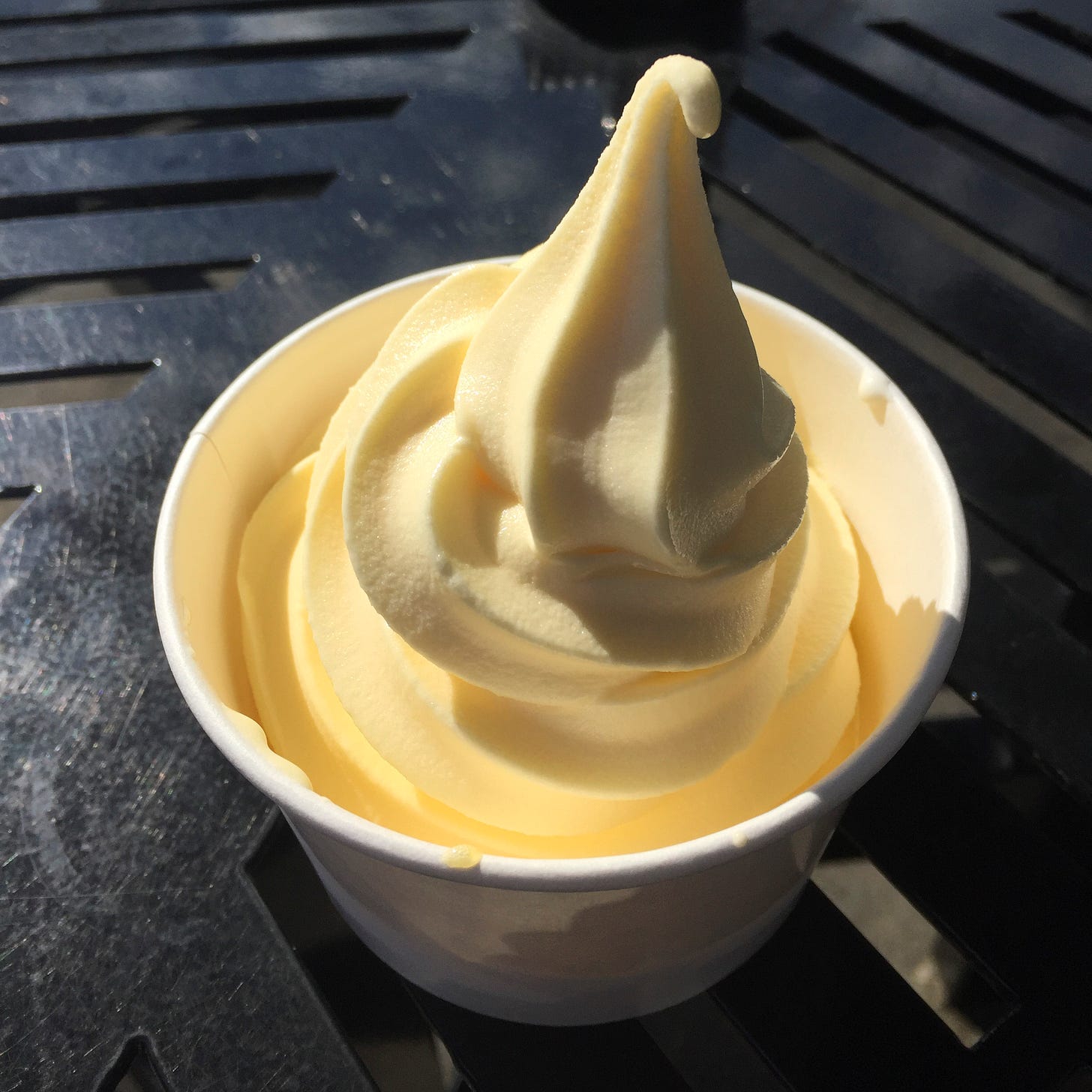 On a black outdoor table in a paper cup, a light yellow swirl of Dole whip.