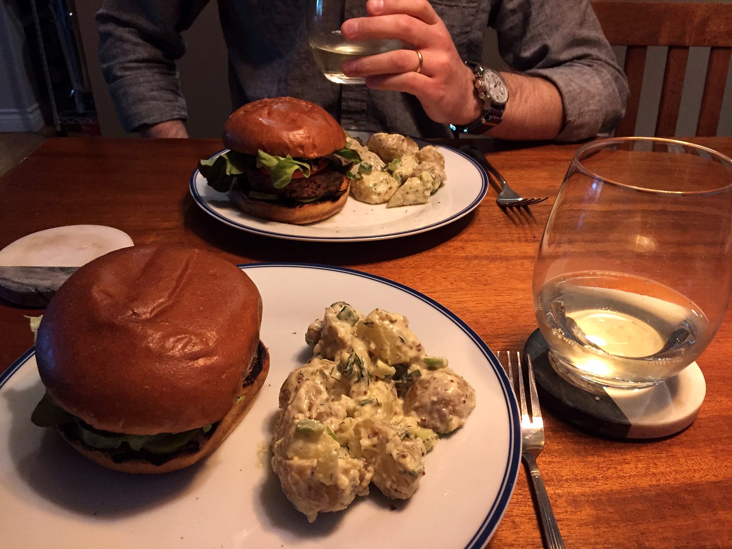 Two white plates across from each other on the table, each with a veggie burger and a serving of potato salad. The potatoes have the skins on and there is lots of dill in the dressing. A glass of white wine sits on a coaster next to the plate in the foreground.