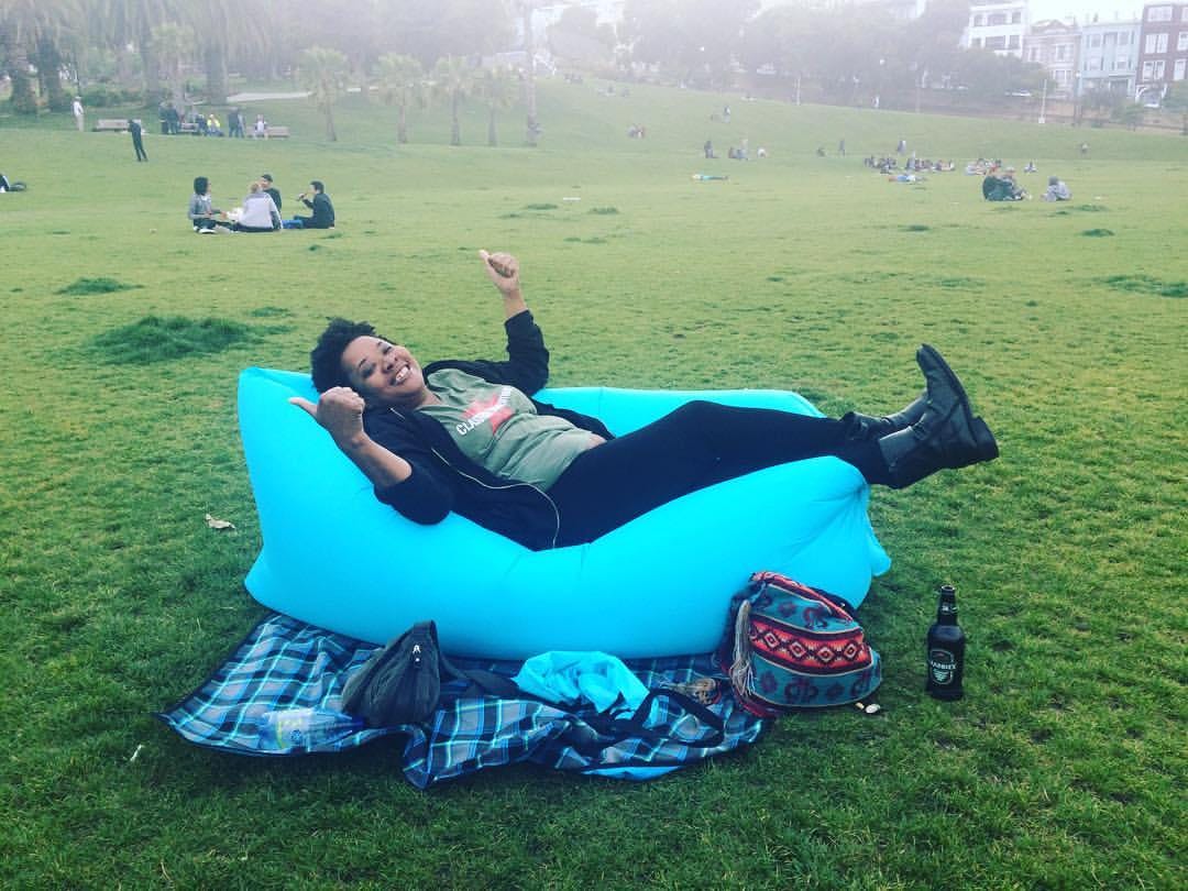 Momos Cheeskos reclining on an inflatable cushion in a park with her thumbs up. She is wearing a green t-shirt and black hoodie with black pants and black boots. She is in a park with a big smile on her face. 