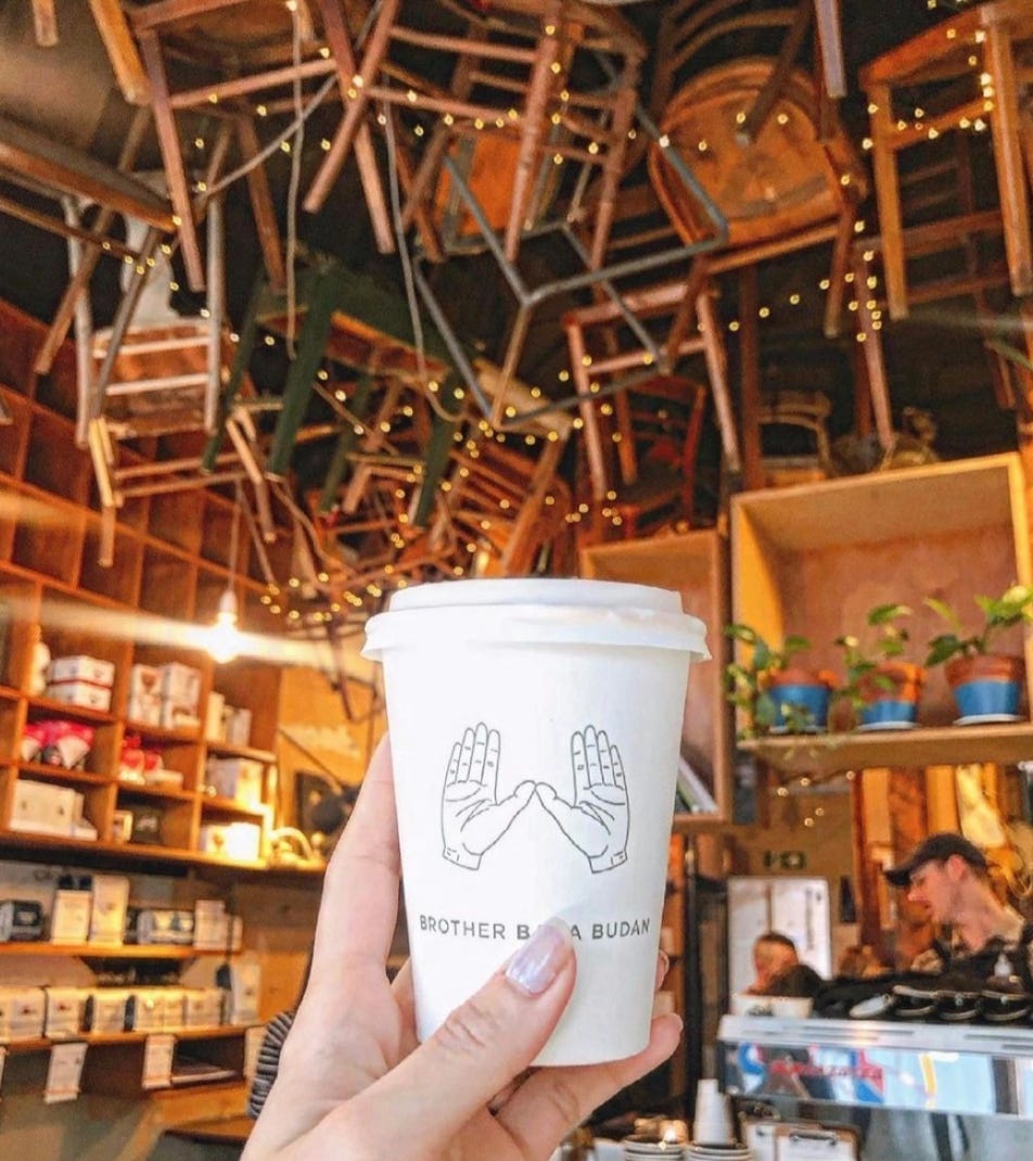 A white to-go coffee cup from Brother Baba Budan cafe is held up in front of the camera. Wooden chairs covered in white string lights hang from the ceiling, and a blurred barista works on a coffee at the bar behind the cup.