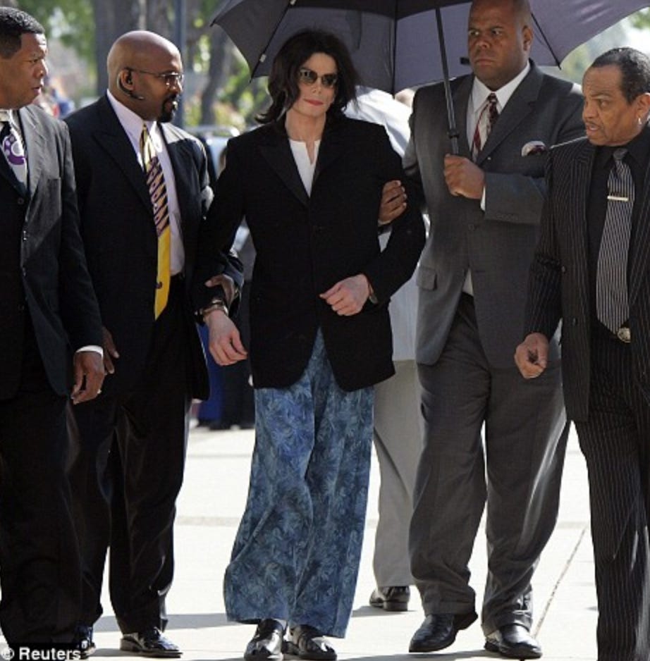 A picture outside of a light-skinned man wearing blue pajama bottoms, a black suit jacket, shoulder length black hair and dark sunglasses. He is supported on either side by darker-skinned Black men holding him up by his arms. One of them is holding an umbrella over his head. There are two other darker-skinned Black men on either side of him on each edge of the picture. All four darker-skinned Black men are wearing dark suits.