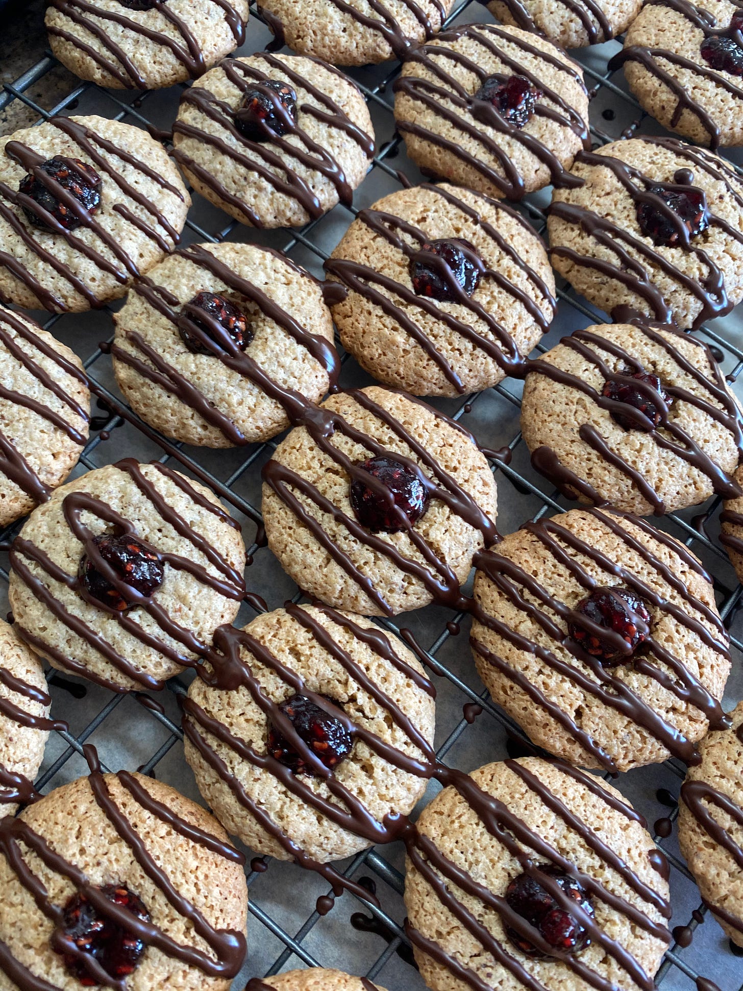 Many pale round cookies sit on a metal cooling rack. They have small dollops of raspberry jam in their centers, and are drizzled with zigzagging lines of chocolate ganache.